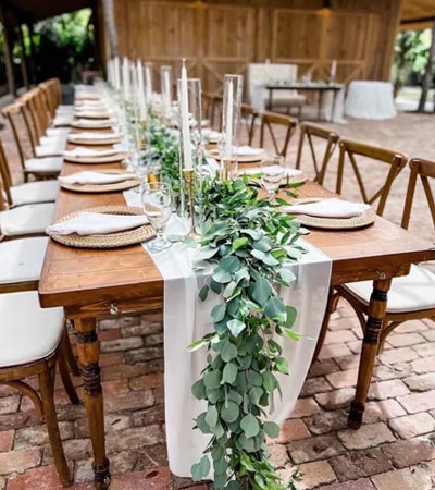 Elegantly styled farm table with a lush eucalyptus garland runner, white linen, woven charger plates, and tall taper candles set on the brick courtyard at The Barn 305, a wedding and event venue in Miami known for its rustic charm, natural greenery, and beautifully curated tablescapes that create an unforgettable dining experience for weddings and private celebrations in South Florida